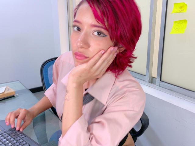 MiaSinclairX with pink hair, pouty lips, and pink shirt, resting her hand on her cheek, sitting at a desk with a keyboard in front of her.