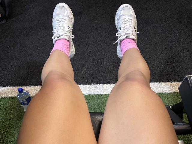 ScarlletZhen's legs, wearing white sneakers, pink socks, and tan skin, with a water bottle on green and black carpet. Her knees are slightly bent.