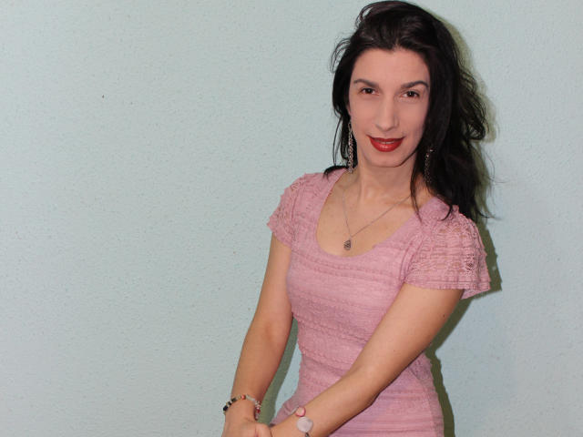 FeelforMe with dark hair, red lipstick, pink lace dress, small tits, necklace, bracelet, smiling, light green wall, standing, hands together, casual, attractive.