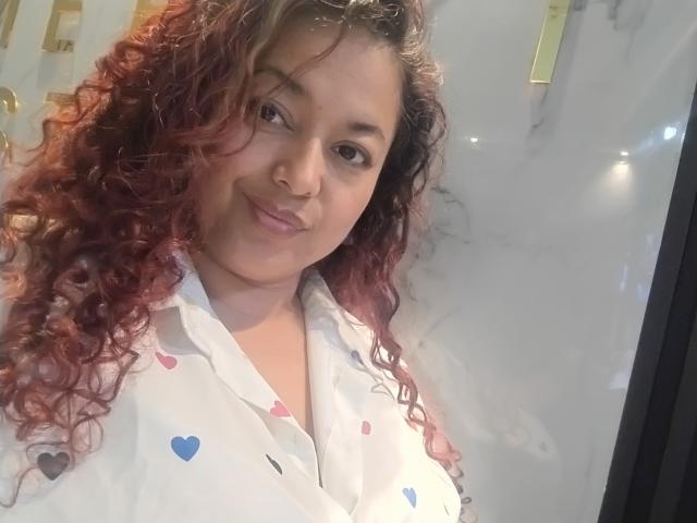 SophiLarusso with curly red hair, smiling, wearing a white shirt with heart patterns, showing her tits, standing in front of a reflective surface, looking at the camera.
