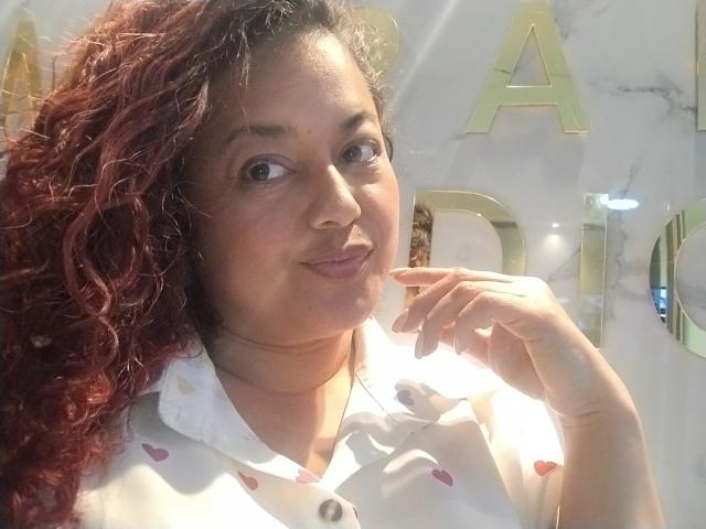 SophiLarusso with curly red hair, medium skin, and small nose, wearing a white shirt with red hearts, smiling slightly, holding her chin, in a well-lit indoor setting.
