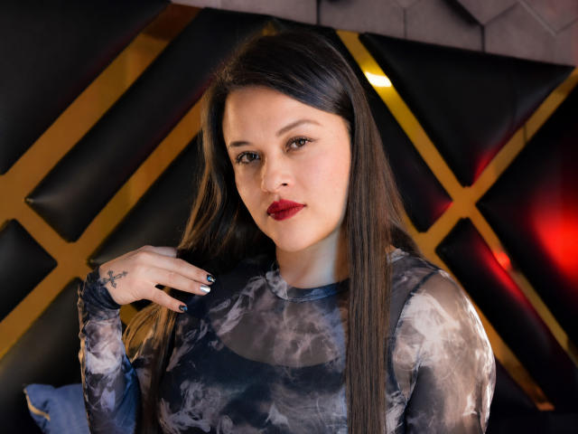 Caroline, avec ses longs cheveux bruns, son rouge à lèvres rouge et son vernis à ongles noir, porte un haut noir et gris tie-dye et a un petit tatouage sur sa main droite. Elle regarde l'appareil photo..