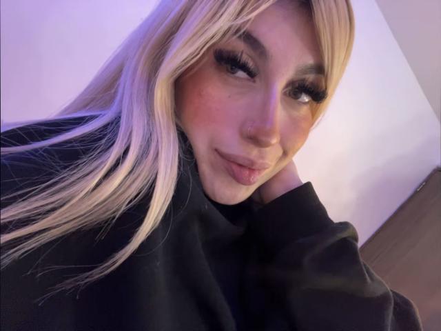 SammyViolet with long blonde hair, dark eyebrows, and thick eyelashes, wearing a black top, head resting on hand, smiling softly, pinkish blush on cheeks, nose ring.