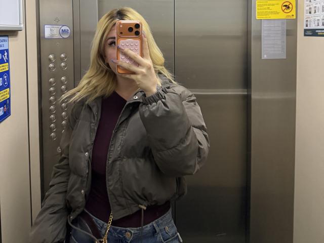 SammyViolet with blonde hair, wearing a puffy green jacket, burgundy top, and blue jeans, taking a mirror selfie in an elevator, phone covering her face.