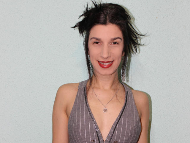 FeelForMe with dark hair, red lipstick, gray pinstriped dress, small tits, necklace, earrings, smiling, standing against pale green wall, slight wind in hair.
