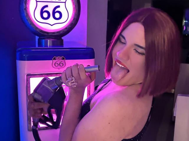 GlossyyBratz with a bob haircut, tongue out, holding a gas pump nozzle to their shoulder, wearing a sparkly black top, under purple and blue lighting. Boobs visible.