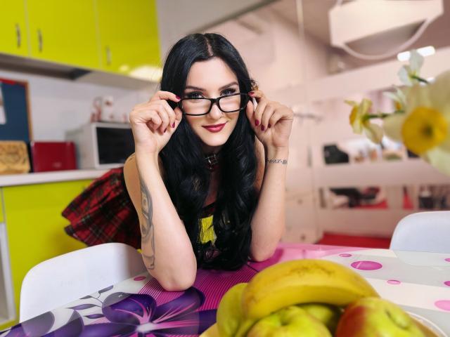 Annabel Rite, met zwart haar, een bril, een rode geruite rok, een gele top, een tatoeage op haar arm, leunt tegen een tafel, glimlacht, houdt een bril vast, en er staan bananen en appels op de voorgrond, in een witte keuken..