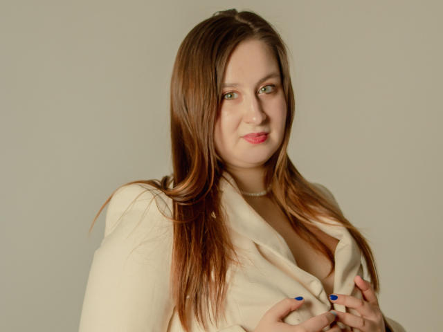 ViaLattea has long brown hair, green eyes, and pink lips. She's wearing a beige shirt, slightly open, showing her tits. She's holding her shirt, with blue nails.