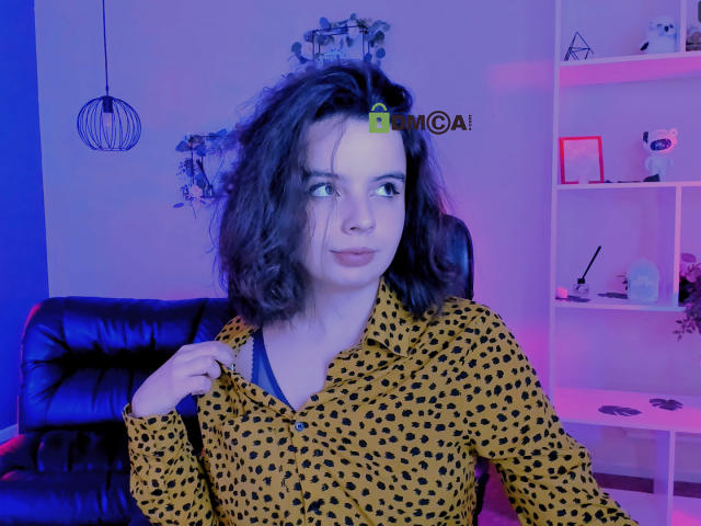 MiamMooon with messy dark hair, wearing a yellow leopard-print shirt, has a hair clip on her head, looking to the side with a slight smile, in a blue-pink lit room.