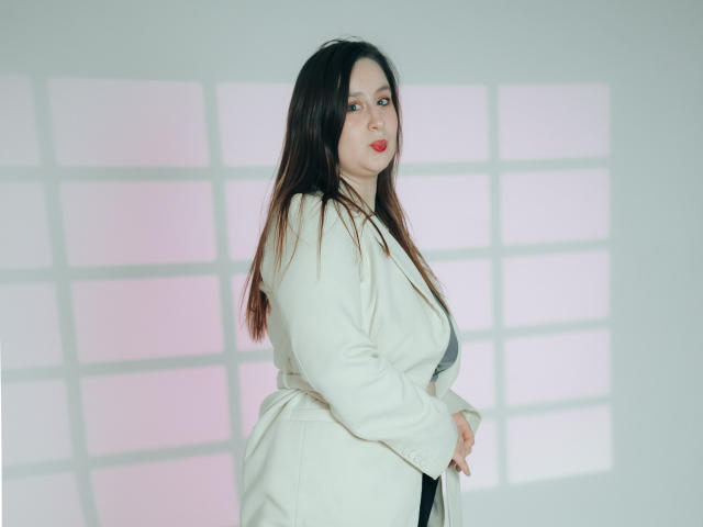 ViaLattea with long brown hair, wearing a white coat, showing her tits, standing sideways, looking at camera with red lipstick. Soft pink and white light in background.