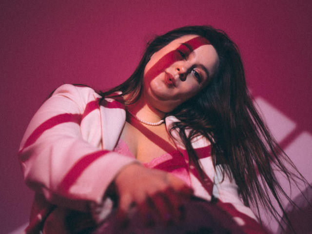 ViaLattea with long brown hair, wearing a pink bra and white striped jacket, shadowed by pink light, looking down with a slightly pouty expression, holding a pink bag.