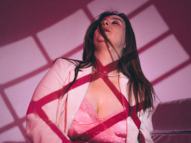 ViaLattea with long brown hair, pink bra, and white jacket, shadowed by red lines, head tilted back, wearing pearl necklace, looking sexy, pink and red lighting.