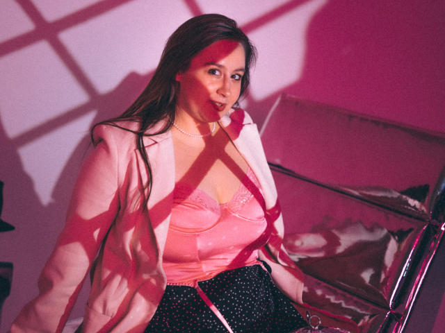 ViaLattea with long brown hair, pink satin bra showing her tits, pink jacket, black polka dot skirt, sitting next to a shiny box, pink and red shadows on her.