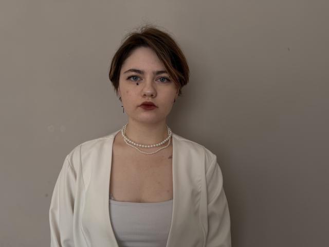 GinnyDelonais has short brown hair, blue eyes, and wears a white shirt with a pearl necklace and open white jacket. Her tits are small, and she has a tattoo.