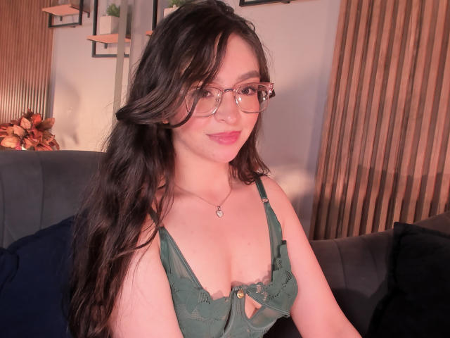 EvaMcAllister mit langem, dunklem Haar, Brille, grüner Schnürung im Dessous-Bereich, die ihre Brüste zeigt, lächelnd, sitzend auf einer dunklen Couch, Holzwand im Hintergrund..