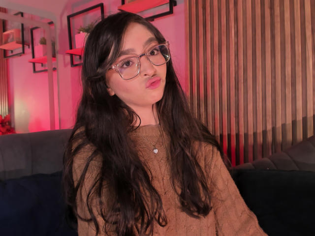 EvaMcAllister met lang zwart haar, die een bril draagt en een bruine trui aanheeft, met haar roze lippen opgetrokken, een hartvormige ketting, zachte roze en rode verlichting, een houten muur, een blauwe bank..