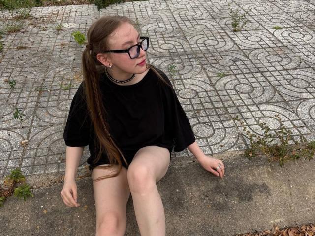 CrystaLux with long brown hair, glasses, black shirt, no pants, sitting on concrete, wearing choker, legs slightly apart, looking up, grass and patterned pavement in background.