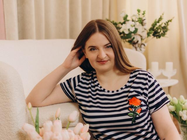 FreyaDak, con i capelli castani, indossa una camicia a righe con un fiore rosso, è seduta su un divano bianco, sorride, ha una mano tra i capelli ed è circondata da tulipani rosa e bianchi..