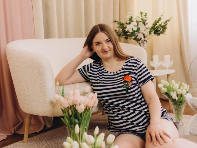 FreyaDak, mit braunen Haaren und einem gestreiften Hemd, sitzt auf dem Boden, die Hand auf dem Kopf, und trägt Jeansshorts. In ihrer Nähe befinden sich rosa Tulpen, ein weißer Stuhl und eine Blumenanordnung. Sie lächelt und sieht niedlich aus..