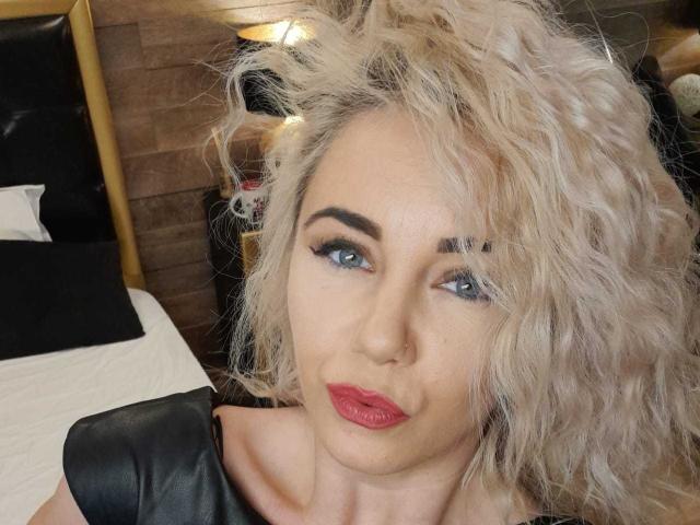 VeraNoires has curly blonde hair, blue eyes, red lipstick, black leather top, and a sexy smile. Her tits are small but she's hot. Her ass is not visible.