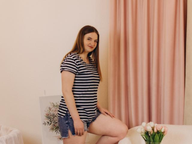 FreyaDak, con i capelli castani, indossa una camicia a righe e pantaloncini di jeans, è seduta su un divano, sorridendo, mostrando il sedere, con dei fiori bianchi in primo piano..