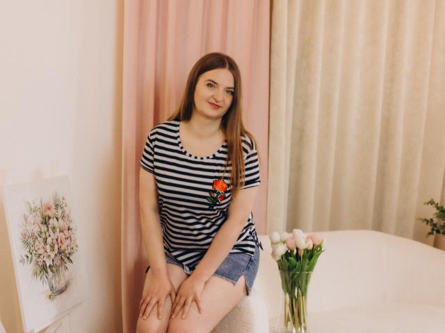 FreyaDak, con la pelle chiara, lunghi capelli castani, indossa una camicia a righe con un fiore rosso e pantaloncini di jeans, seduta, sorridente, con le mani sulle ginocchia, dietro di lei tende rosa e beige..