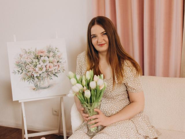 FreyaDak, com cabelo castanho, sorrindo, vestindo um vestido branco com bolinhas, segurando um vaso de vidro com tulipas brancas, sentada em um sofá bege, ao lado de uma pintura floral..