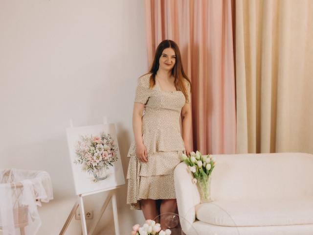 FreyaDak with brown hair, wearing a beige polka-dot dress, standing next to a flower painting and white sofa, holding a vase of white tulips.