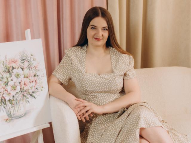 FreyaDak, avec de longs cheveux bruns, portant une robe beige à pois, assise sur un canapé beige, souriante, les mains posées sur ses genoux, à côté d'un tableau représentant un bouquet de fleurs..