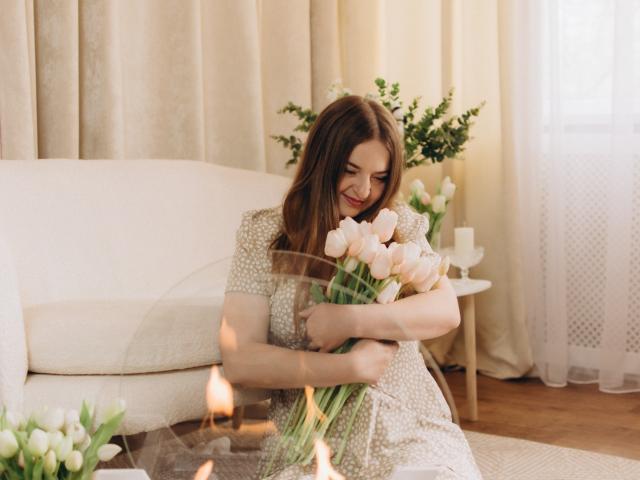 FreyaDak with brown hair, smiling, hugging a bouquet of pink tulips, wearing a white floral dress, sitting on a beige couch, candles in the foreground.