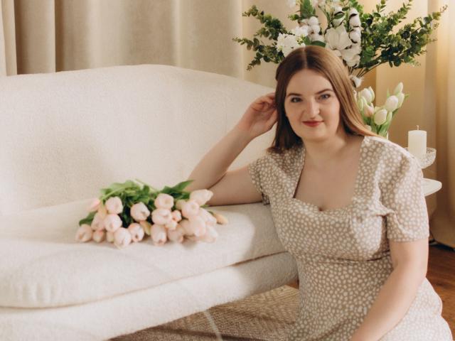FreyaDak with brown hair, wearing a polka-dot dress, sitting on a white couch with pink tulips, arm on the back, small tits, cute smile, floral vase in the background.