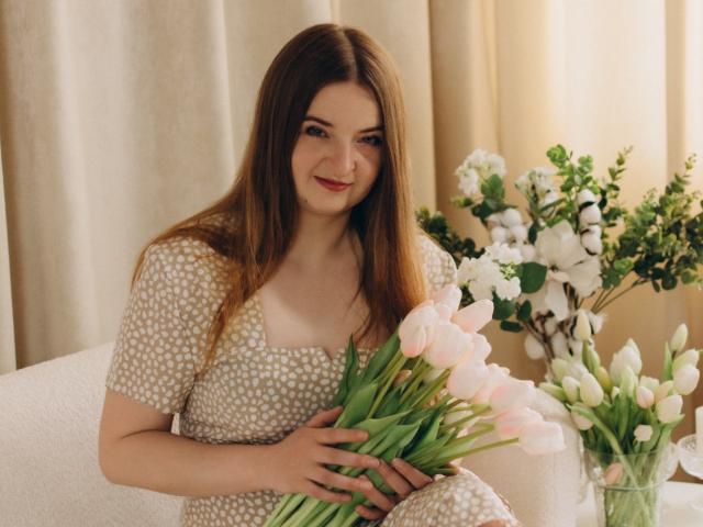 FreyaDak mit langen, braunen Haaren, die ein weißes, gemustertes Kleid trägt, rosa Tulpen hält und auf einem weißen Stuhl sitzt, lächelt sanft. Im Hintergrund steht eine weiße Blumenvase..