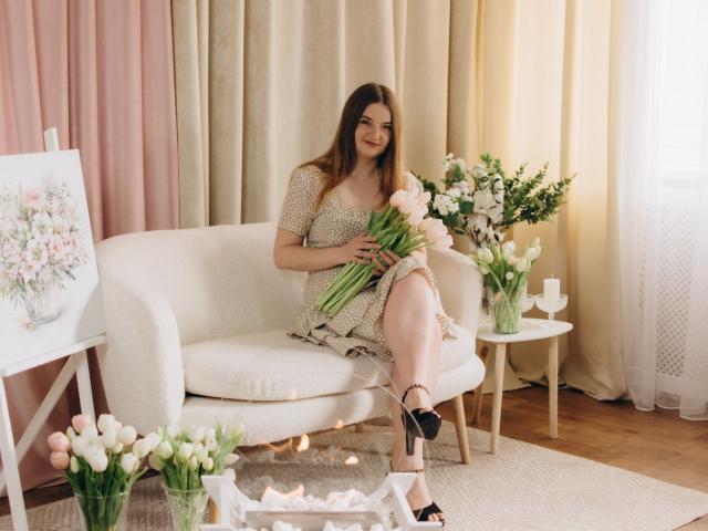 FreyaDak, avec de longs cheveux bruns, est assise sur un canapé blanc, tenant des tulipes roses, vêtue d'une robe beige, de talons noirs, et elle sourit. Une décoration florale l'entoure..