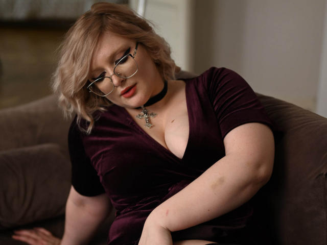 CoolTatis with blonde, wavy hair, glasses, red lipstick, and a black choker, sitting on a couch in a purple velvet dress, showing her tits and a bruise on her arm.
