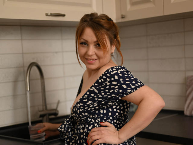 AnitaLevantaa, with brown hair in a messy bun, smiling, wearing a navy blue dress with white patterns, standing in a kitchen, hand on hip, showing her tits.