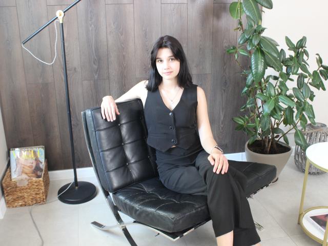 LettyCurly with black hair, wearing a black vest and pants, sits on a black leather chair, her ass on the seat, arms resting, legs crossed, in a modern room.