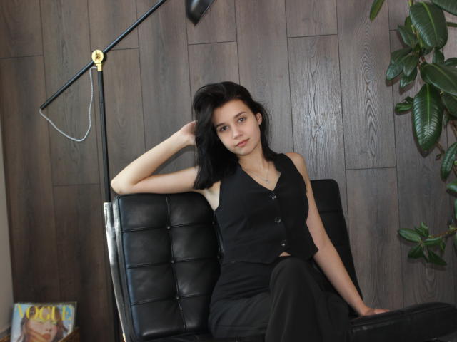 LettyCurly with black hair, wearing a black button-up top and black pants, sits on a black leather chair, hand in hair, lamp above, green plant to the right.