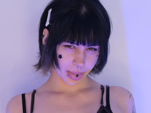 AshleyAlt has black hair, star face tattoos, piercings in nose and tongue, and a white hair clip. She's wearing a black strap top, with a blue-purple light.