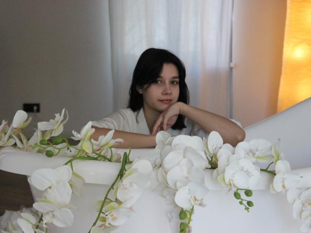 LettyCurly with black hair, white shirt, leaning on white table, looking at camera, surrounded by white orchids, soft light from right, relaxed expression, hands near chin.