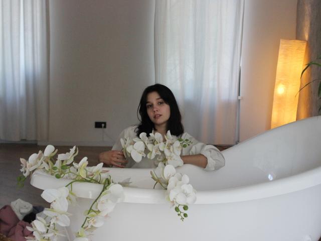 LettyCurly with dark hair, white blouse, sitting in a white bathtub, surrounded by white flowers, looking at camera, soft yellow lamp on right.