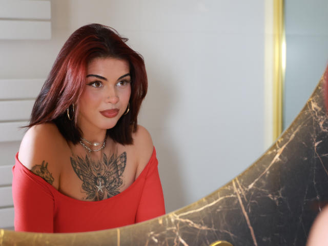 NovaSin with red hair, wearing an off-shoulder red top, showing a large butterfly tattoo on her tits, looking in a mirror, smiling slightly, gold necklace, white wall.