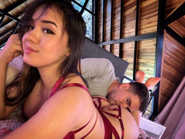 HannaAndNico, avec des cheveux foncés, en lingerie rouge à bretelles fines et avec de petites seins, allongées sur le lit, la tête dans la main, la tête de Nico sur ses fesses, pièce avec des panneaux de bois, souriantes, yeux marrons, mignonnes..