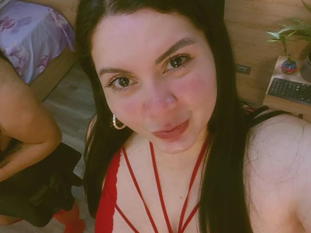 RachelCarlton with long black hair, red strappy top, and gold hoop earrings, looking up at the camera with a slight smile, showing her boobs. Another person's ass is visible.