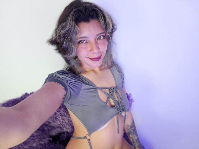 DarckVelvet with grayish-brown hair, wearing a gray, strappy top that exposes her tits, smiling at the camera, has a nose ring, and a tattoo on her left arm.