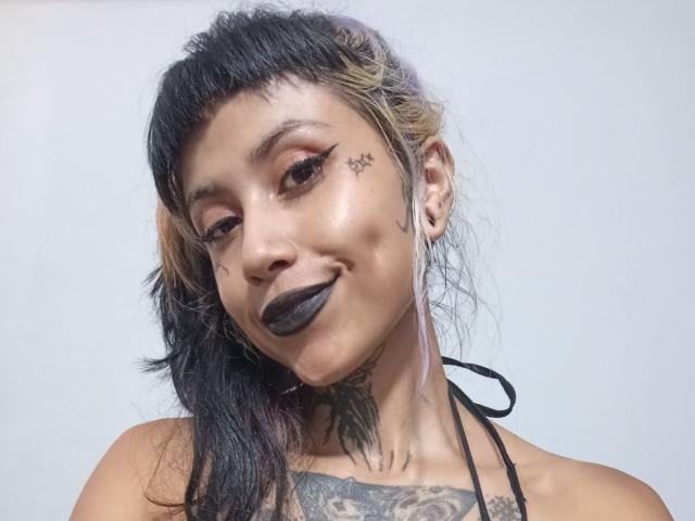 NyxVega with black lipstick, dark eyeliner, and a star tattoo on her cheek, smiling. She has black and blonde hair, tattoos on her neck, and ear plugs.