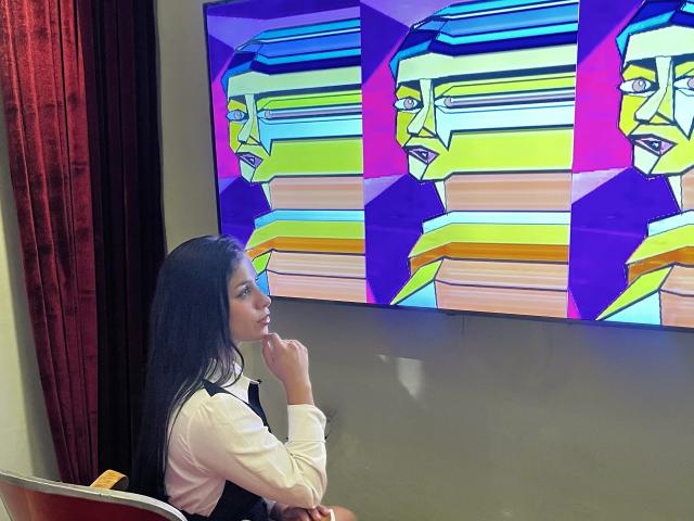 MelodyRoss with long black hair, white shirt, and black vest, looks at colorful, abstract paintings of faces on a wall. Her finger rests on her chin, looking curious.