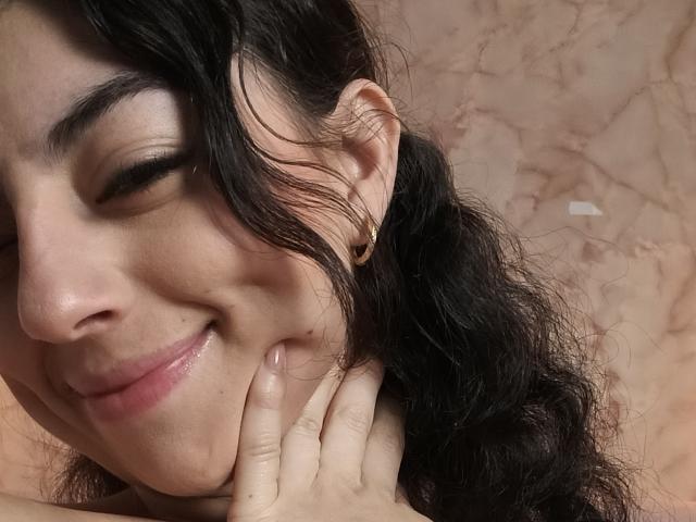 AnntonellaWatson with black curly hair, eyes closed, smiling, holding her cheek with a finger, wearing a small gold earring, pinkish skin, and glossy lips.
