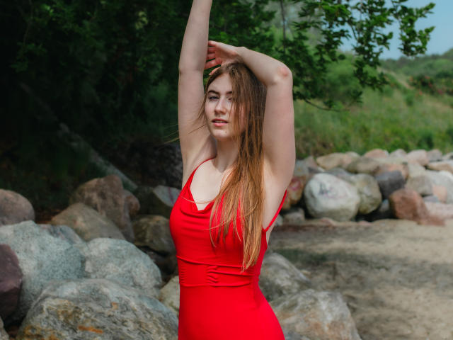 MeganCandy with long red hair, arms raised, wearing a red dress, standing among rocks, smiling, showing her tits, in a forested area.