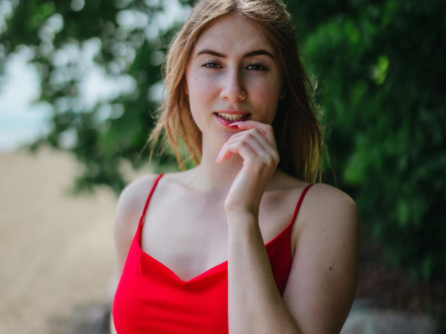 MeganCandy with blonde hair, red tank top, fair skin, and a cute smile, touching her lip with her finger, showing her tits. Green foliage blurred in the background.