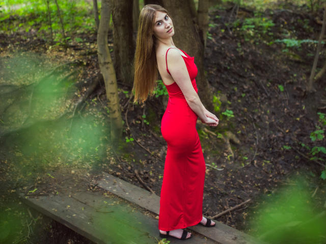 MeganCandy in a red dress, standing on a wooden path, looking over her shoulder, showing her ass, with long brown hair, wearing black sandals, in a forest.
